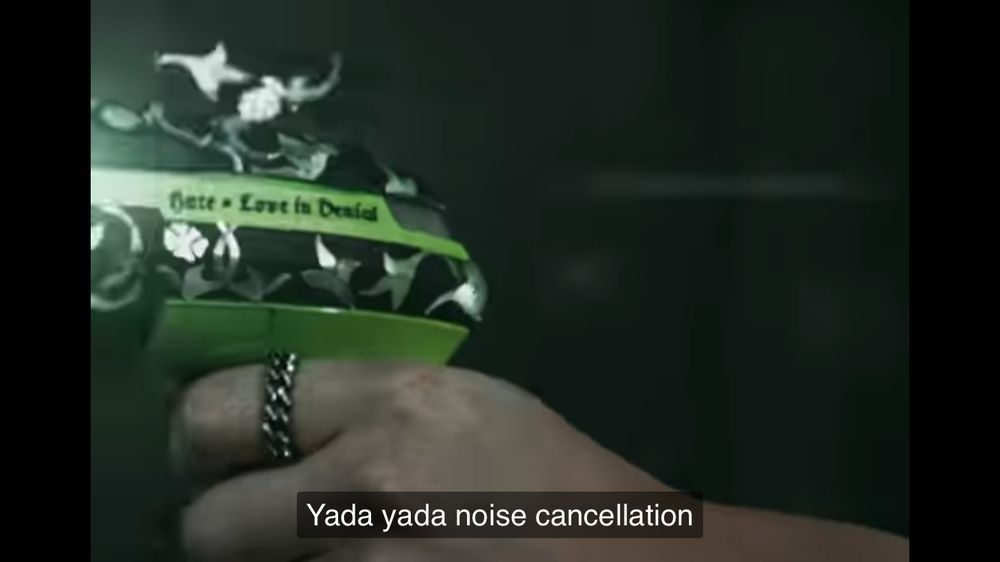 A screenshot from the Stray Kids BLEEP Unveil video with the side of a bright green scan gun held in a hand. The text on the gun says “Hate = Love in Denial”. The video subtitle reads “yada yada noise cancellation”.