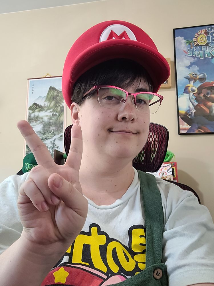 A selfie of redria in green overalls and wearing a mario hat, giving a peace sign
