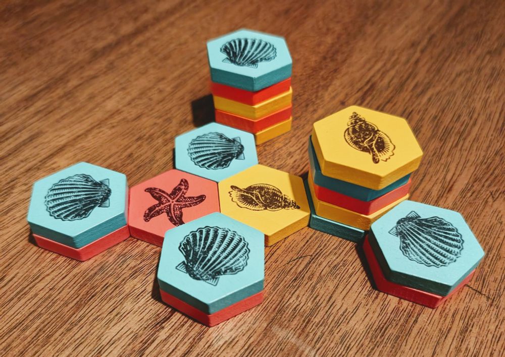 A photo of High Tide. Pink, cream, and blue hexagonal tokens are arranged and stacked on a wooden table. Each token has a screen printed seashell on it.