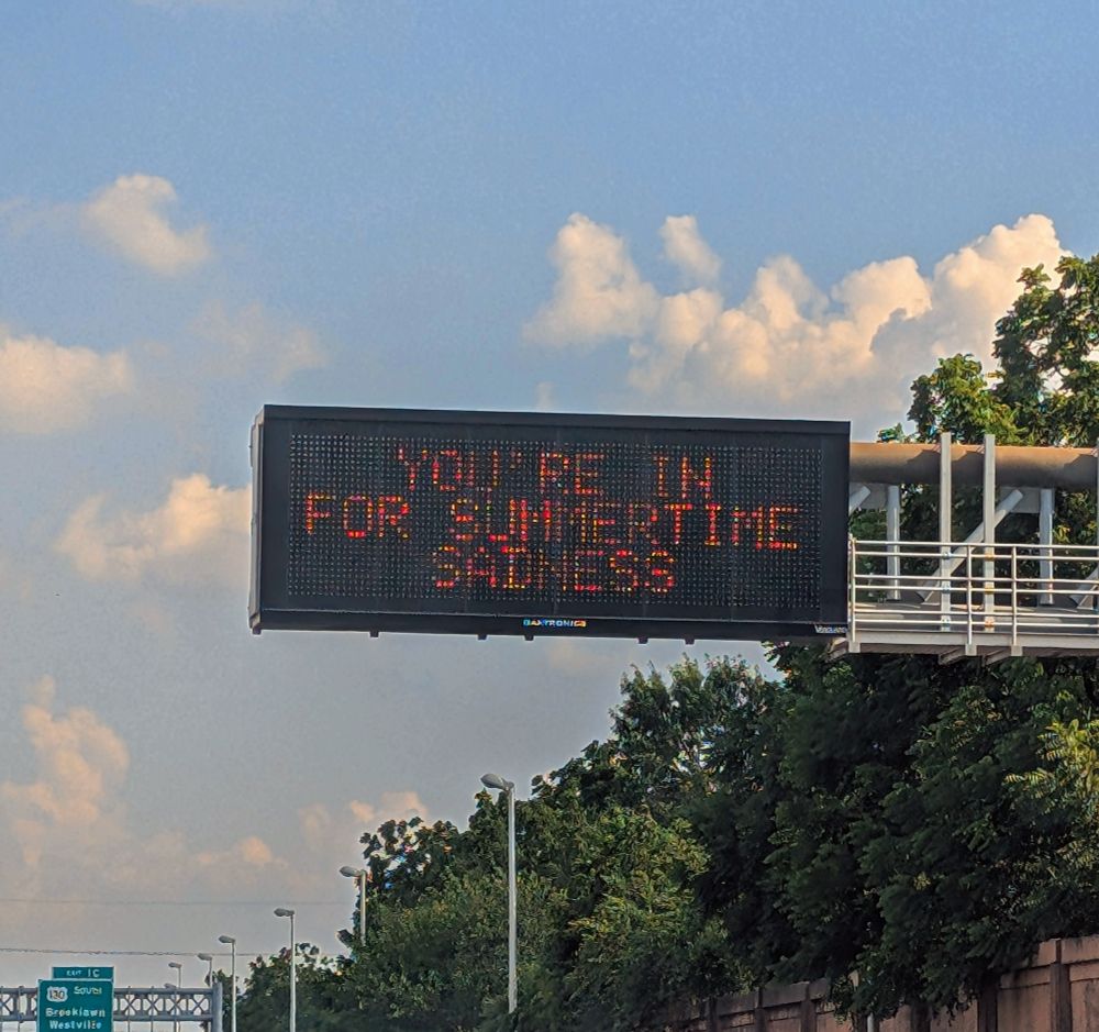 A photo of an LED info sign on the highway. It reads "You're in for summertime sadness" in all caps. Some trees, highway walls, and sky surround the sign.