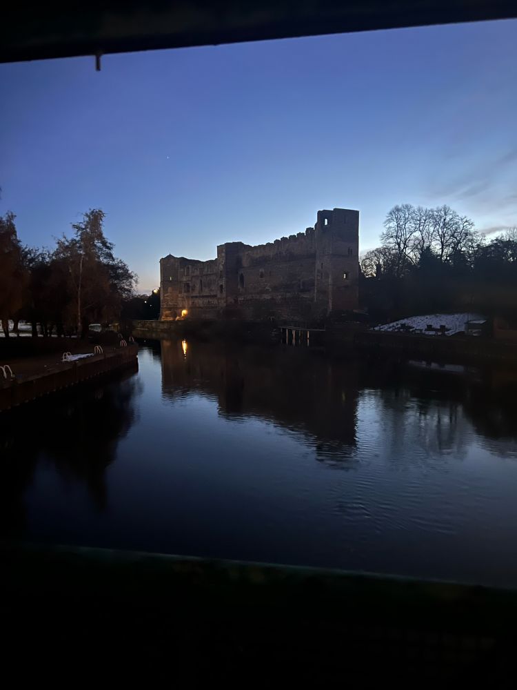 Photograph of a castle on the river in England in the early morning light. The castle is reflected in the water and there is lingering snow on the banks beside it. 