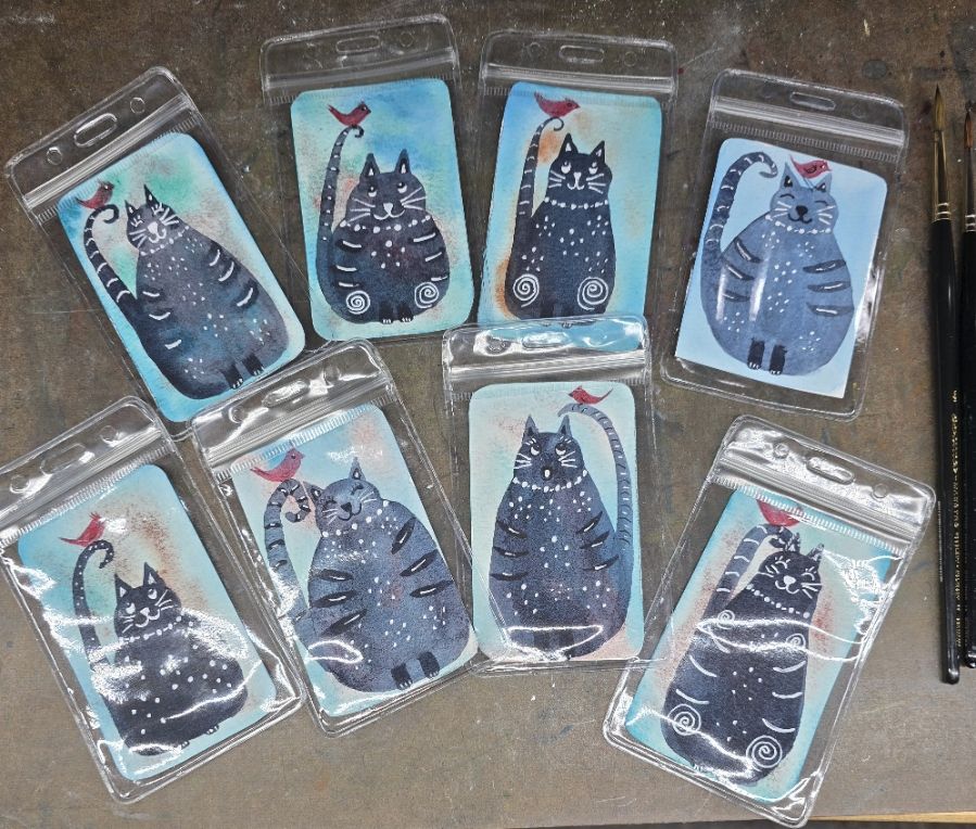 A photo of 8 clear luggage tags. Inside each is a small watercolor painting of a whimsical, dark blue cat with a red bird. The cats are embellished with white dots and lines. 