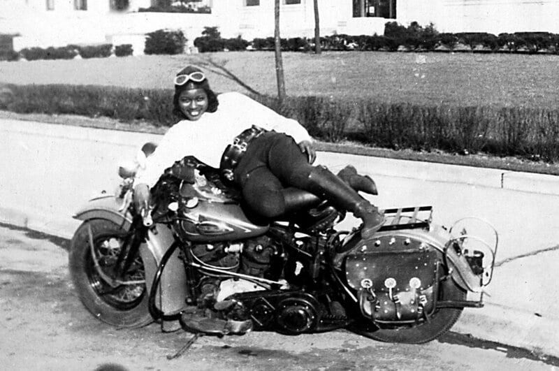 A black and white photo of a young Black woman lying atop an old-timey Harley-Davidson motorcycle. She's wearing knee-high riding boots, jodphurs, a black leather kidney belt, a white long-sleeved top, a leather Snoopy helmet, and goggles. She has a huge smile on her face and he holding something (a beer can, maybe?) in her right hand. 