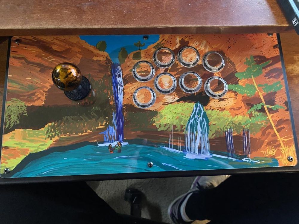 A fight stick with a picture of a pair of waterfalls going down a cliff into a pond at the base. In the pond and at the base of the taller waterfall, two women practice with wooden swords. Art is OC and I also designed and assembled the fightstick from parts. Painting made in Corel Paint