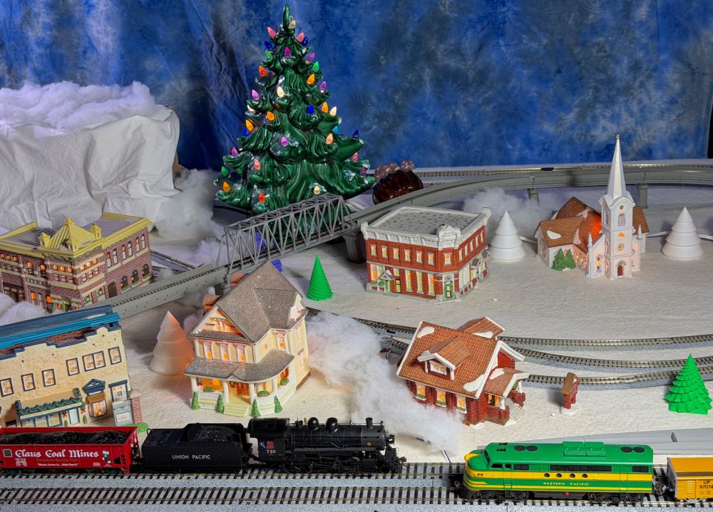Model railroad scene. Two locos are heading towards each other on a double track. A black steam loco with a red "Claus Coal Mines" coal car is heading to the right and the John Deere colored (it's actually Western Pacific) diesel loco is headed east. 

Behind them are some of the buildings from the Christmas in Lindsborg set of ceramic prominent buildings from town.

My grandma's pottery Christmas tree, from over 50 years ago, is at t he back.
