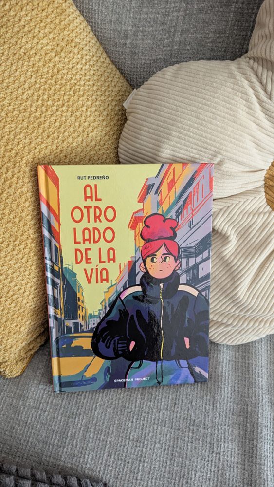 Cómic "el otro lado de la vía" posado entre dos cojines. En la portada aparece el dibujo de una chica con sudadera y moño con la ciudad de fondo.