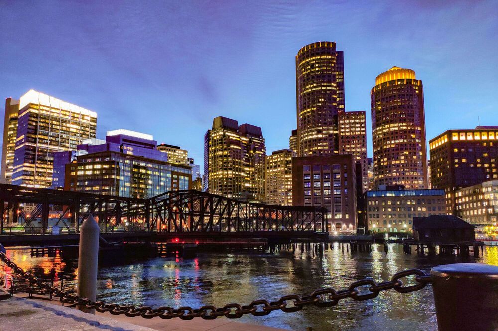 Image of downtown boston with water in the forefront and buildings in the background. Image taken in evening with the sun mostly set.