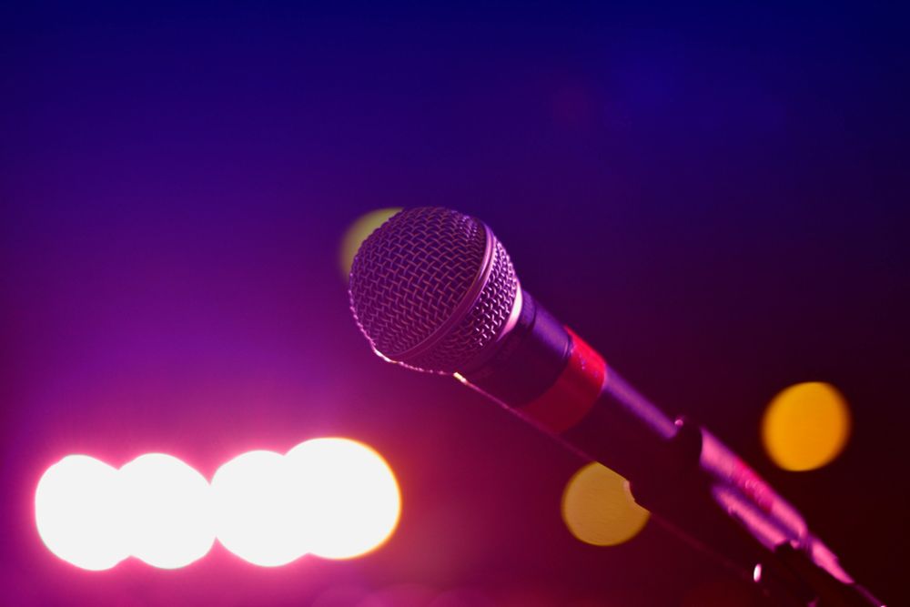 Close-up of a microphone on a stand with colorful stage lights in the background, featuring vibrant pink, purple, and blue tones, suggesting a performance or concert setting.