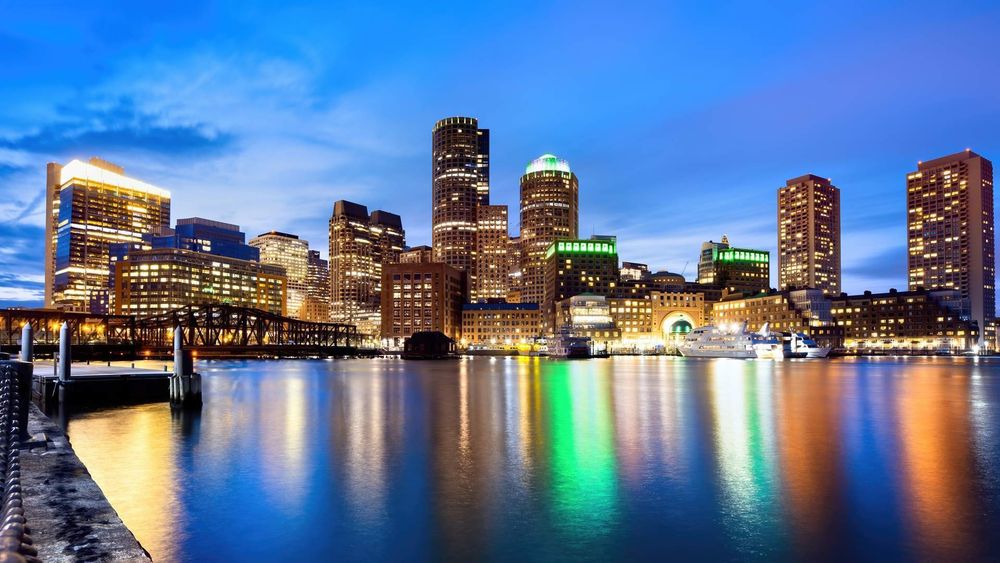 A vibrant view of the Boston skyline at dusk, with illuminated skyscrapers reflecting in the calm waters of the harbor. A dock and moored boats are visible in the foreground, while the sky transitions from deep blue to soft twilight hues.