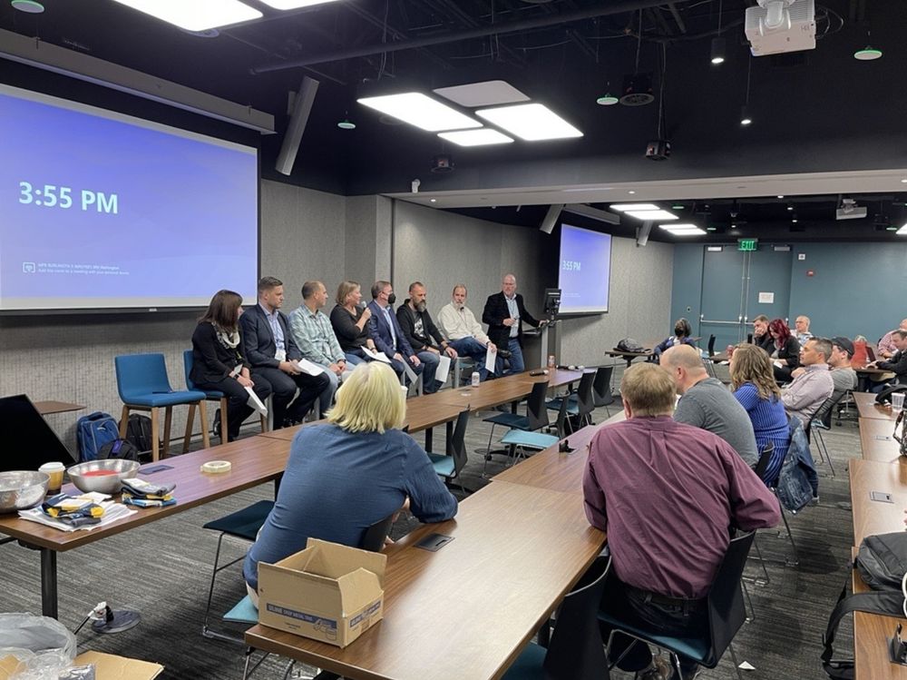 A panel discussion at CollabDays New England shows nine speakers seated in a row at the front of a conference room, with one person standing at a podium speaking. The large projection screen behind them displays the time "3:55 PM." Attendees are seated at tables facing the panel, engaged in the session.