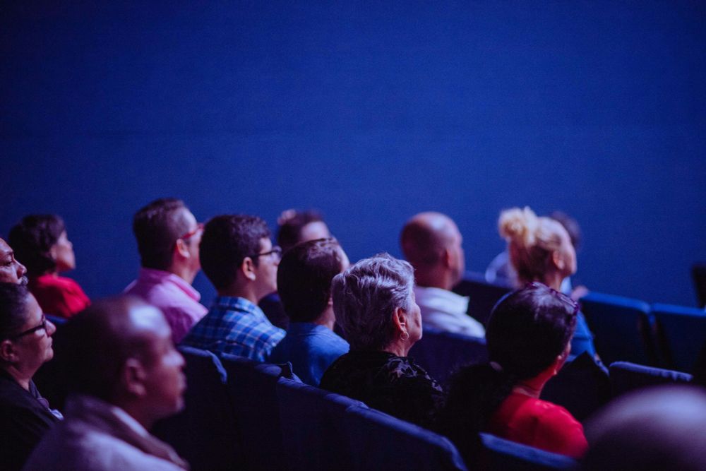 Audience members seated in rows, attentively facing forward in a dimly lit auditorium with a blue backdrop. The group is diverse in age and appearance, and the lighting creates a focused, contemplative atmosphere.