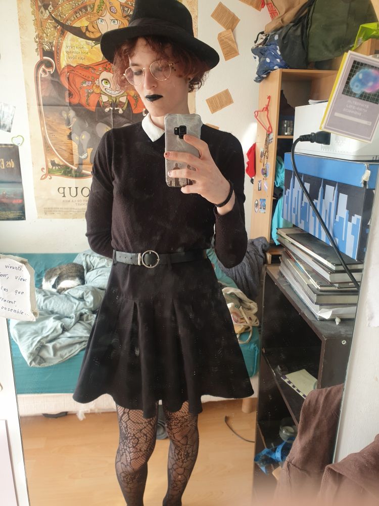 Zélie, une personne avec une jupe joire, un pull noir avec un coll claudette et des collants toile d'araignée. Il a du noirà lèvres et un chapeau, notamment