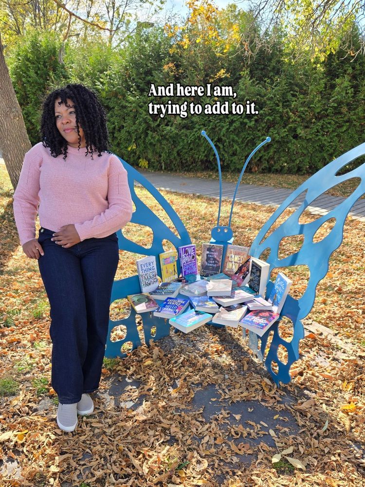 Roxanne LoMarc, author of Love And Other Two-Player Games, stands beside a large, blue, metal butterfly seat that is loaded with books from her newsletter giveaway stash. The text reads, "And here I am trying to add to it."