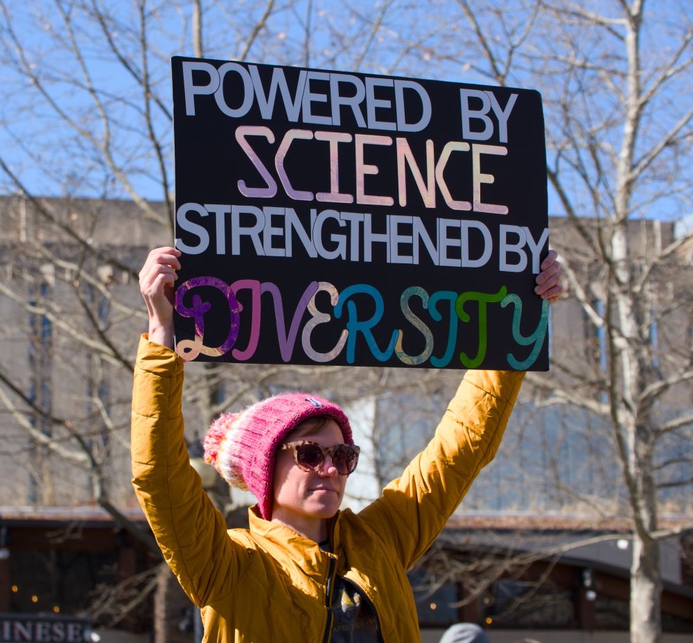 Person in pink hat holding sign saying "Powered by science strengthened by diversity" with diversity in reflective colored letters from purple to green. 