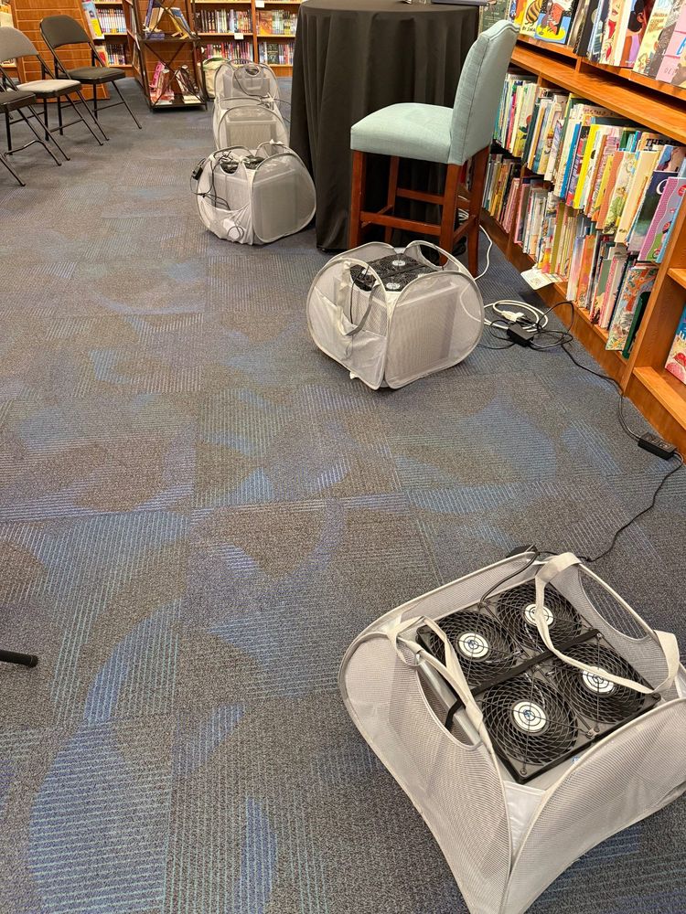 Five AirFanta 3Pro air filters in mesh bags on the floor near a table and chair at the front of a room. Lots of books on shelves all around. 