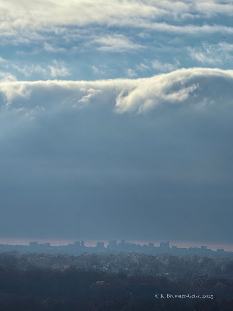 Photo of a city skyline silhouette against a pale red bit of sky. Above the city is a huge cloud formation that looks like a breaking ocean wave. Above the cloud is an airplane in blue sky with white clouds heading out of the photo. 