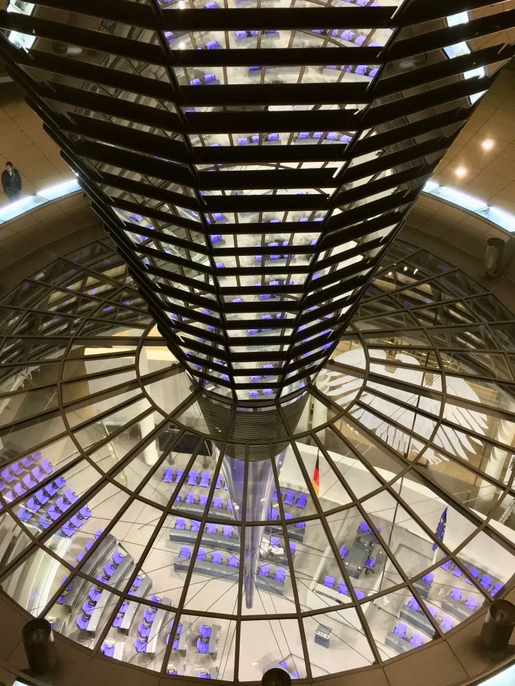 Das Bild zeigt eine Innenansicht eines modernen Gebäudes mit einer großen, gläsernen Kuppel. Zentral im Vordergrund befindet sich ein spiralförmiges, verspiegeltes Element, das sich von oben nach unten erstreckt und als zentrales architektonisches Merkmal wirkt. Durch die gläsernen Strukturen sind darunter viele Reihen lilafarbener Stühle sichtbar, die in einem halbrunden Muster angeordnet sind. Die Sitzordnung deutet auf einen Plenarsaal hin. Auf dem Boden ist das stilisierte Abbild eines Adlers zu erkennen. Im Hintergrund sind Fahnen zu sehen, darunter die deutsche und die europäische Flagge. Das Bild ist aus einer erhöhten Perspektive aufgenommen, sodass die Symmetrie des Raumes und die Architekturelemente gut zur Geltung kommen. Es sind keine Menschen im Hauptbereich zu sehen, nur vereinzelt am Rand.