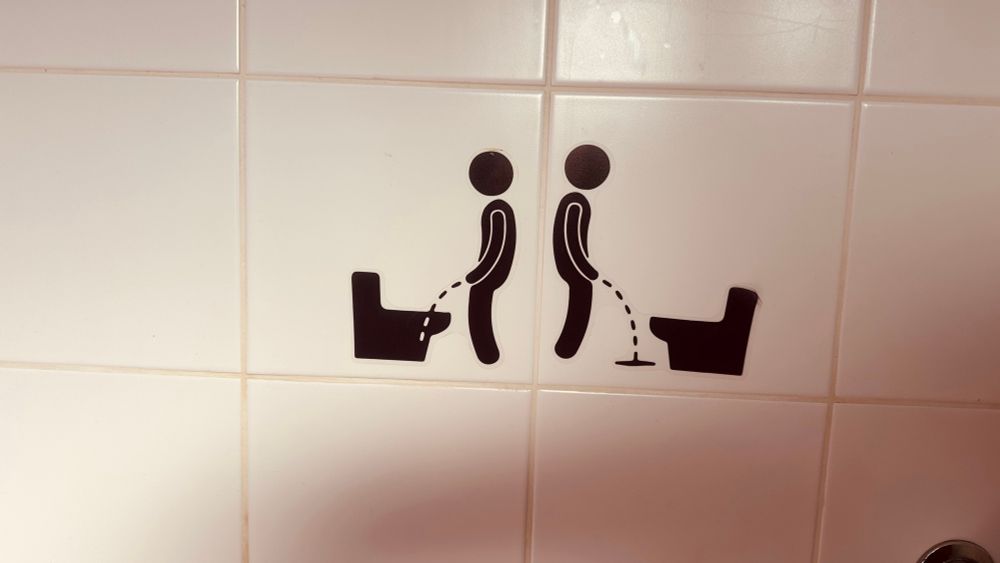 Das Foto zeigt zwei schwarz-weiße Piktogramme auf weißen Fliesen. Beide Bilder stellen stilisierte Figuren dar, die vor einer Toilette stehen. Links ist eine Person zu sehen, deren Urinstrahl in die Toilettenschüssel führt. Rechts steht ebenfalls eine Person vor einer Toilette, deren Urinstrahl jedoch auf den Boden neben die Schüssel trifft. Die Motive sind klar voneinander abgegrenzt und ohne weitere Symbole oder Text dargestellt. Die Fliesen sind schlicht und weisen keine weiteren Verzierungen auf. Die Bildaussage konzentriert sich rein auf die dargestellte Handlung der Figuren.