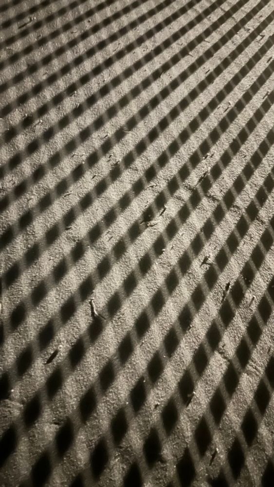 Das Bild zeigt eine gleichmäßige Schattenstruktur auf einer leicht körnigen Fläche. Die Schatten verlaufen diagonal von der oberen linken zur unteren rechten Ecke und bilden ein regelmäßiges Muster aus parallelen, rautenförmigen Feldern. Die Lichtquelle ist offensichtlich stark und wirft harte, klare Schatten, vermutlich durch ein Gitter oder eine andere perforierte Oberfläche. Die Oberfläche darunter weist leichte Unebenheiten und kleine, verstreute Fragmente auf. Die Farben sind in gedeckten Tönen gehalten, überwiegend Grau- und Beigenuancen. Das Gesamtbild wirkt abstrakt und betont das Muster aus Licht und Schatten, das sich gleichmäßig über die Fläche zieht.