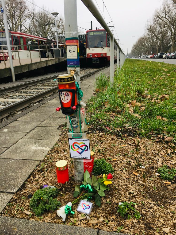 Das Foto zeigt eine Straßenbahnhaltestelle an einem trüben Tag. Im Vordergrund befindet sich ein Gedenkort mit Blumen, Kerzen und mehreren bunten Schildern mit einem Herzsymbol. Auf einem Schild steht: „Kiterliebe ist unendlich!“ Daneben liegen Schnittblumen, Pflanzen und weitere Kerzen. Der Gedenkort ist an einem Mast direkt neben den Schienen angelegt. Im Hintergrund sieht man eine wartende Straßenbahn der Linie U79 sowie wartende Fahrgäste am Bahnsteig. Links verläuft der Bahnsteig, rechts wächst Gras und Bäume säumen die Straße. Im Hintergrund ist eine lange Reihe parkender Autos zu erkennen.