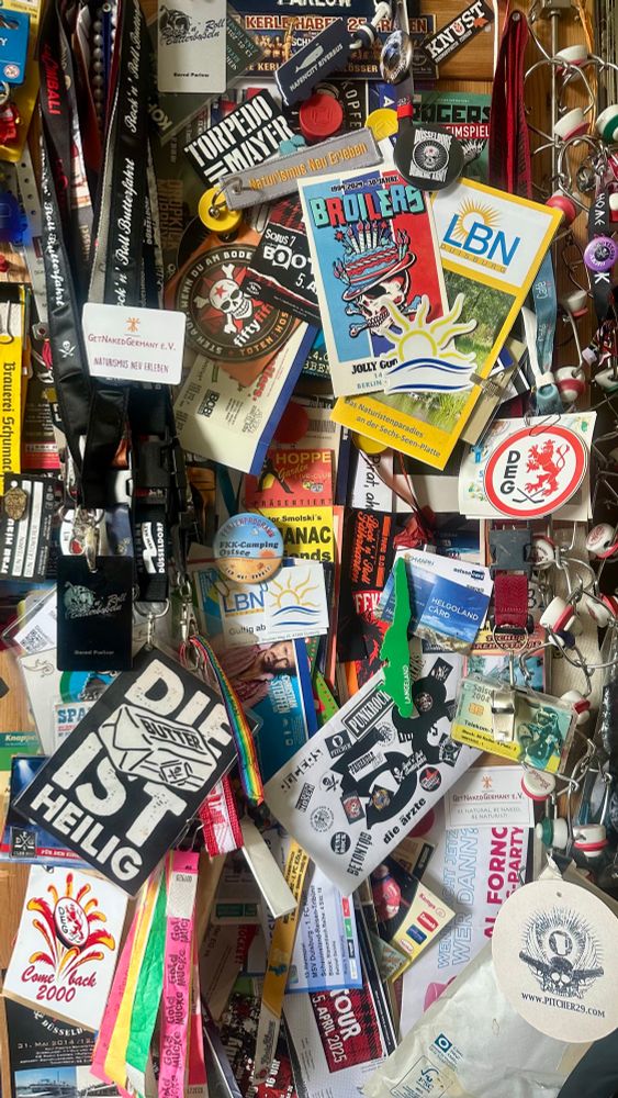 Das Bild zeigt eine Pinnwand, die dicht mit Eintrittskarten, Ausweisen, Festivalbändchen und diversen Sticker bespickt ist. Im Mittelpunkt hängen mehrere bunte Karten und Ausweise mit auffälligen Schriftzügen, wie “FKK-Camping Ostsee”, “BROILERS”, und LBN. Neben den Karten sind zahlreiche Schlüsselbänder und Armbänder befestigt, die unterschiedliche Farben und Logos tragen. Es sind verschiedene Veranstaltungsflyer, Kühlschrank-Magnete und Tickets zu sehen, wie beispielsweise „Rock’n’Roll Butterfahrt“, “Torpedo Mayer”, “Pitcher29.com” und “Naturismus neu erleben”. Die Gegenstände sind eng aneinandergereiht und übereinandergelegt, wodurch ein vielschichtiges und lebendiges Gesamtbild entsteht. Es sind auch Vereinslogos und Sticker mit Motiven oder Texten vertreten. Die Anordnung wirkt zufällig, die einzelnen Elemente überdecken sich und ragen mehrfach über den Rand der Pinnwand hinaus.