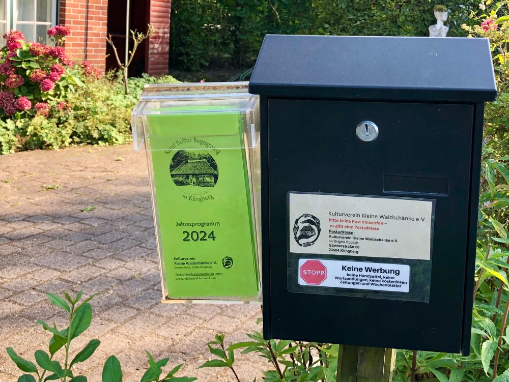 Das Bild zeigt einen Gehweg neben einem Garten mit blühenden Hortensien und einer rot verklinkerten Hausfassade im Hintergrund. Im Vordergrund stehen zwei Kästen an einem Holzpfosten: Links befindet sich ein transparenter Kasten mit einem grünen Faltblatt, das die Aufschrift „Jahresprogramm 2024, Kunst Kultur Begegnung in Klingberg“ trägt. Rechts daneben ist ein schwarzer Briefkasten mit zwei Aufklebern; einer enthält Hinweise zum Kulturverein Kleine Waldschänke e.V. mit Postadresse und der Anmerkung, keine Post einzuwerfen. Der andere Aufkleber signalisiert „Keine Werbung – keine Handzettel, keine Wurfsendungen, keine kostenlosen Zeitungen und Wochenblätter“ mit einem roten Stoppschild-Symbol. Im Hintergrund sind Gartenpflanzen und eine weiße Figur zu erkennen.