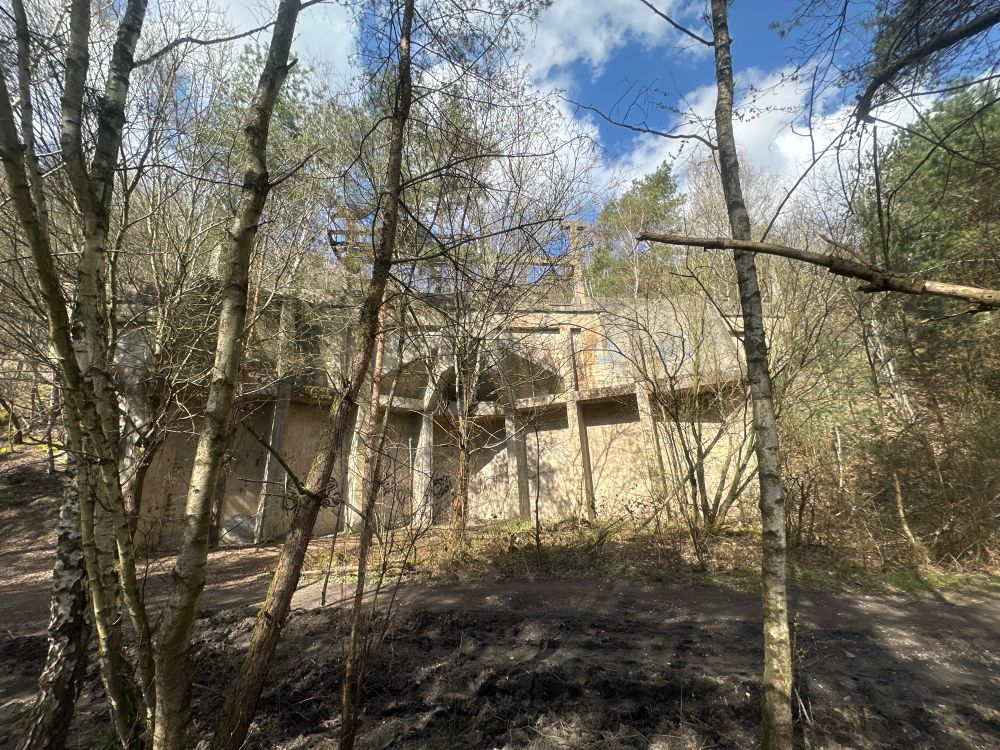 Das Bild zeigt eine größere, verlassene Betonstruktur, die teils von Bäumen und Gebüsch umgeben ist. Das Gebäude befindet sich mitten im Wald, die Fassade ist von mehreren Säulen geprägt und teils von Graffiti bedeckt. Die Szenerie ist von unbelaubten, dünnen Baumstämmen im Vordergrund dominiert. Der Boden wirkt uneben und stellenweise matschig, während im Hintergrund teilweise blauer Himmel und weiße Wolken sichtbar sind. Die gesamte Atmosphäre ist ruhig und von natürlicher Vegetation geprägt.