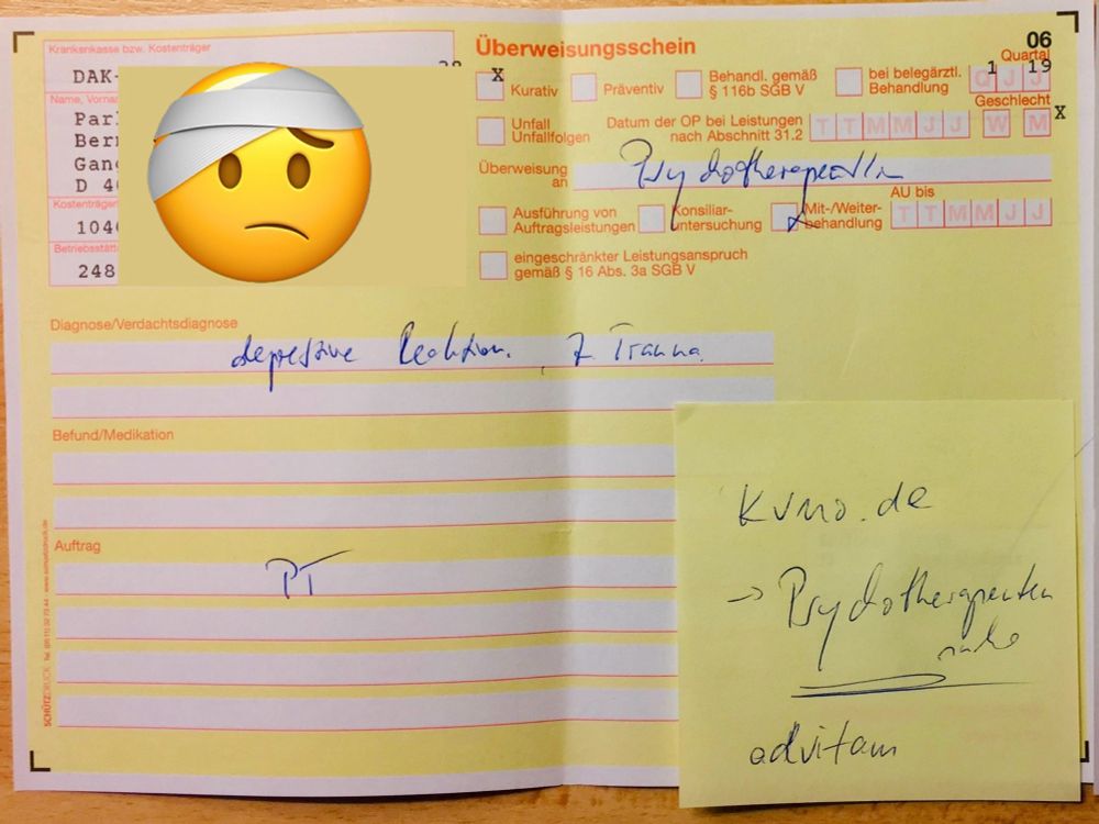 Auf dem Bild ist ein gelber Überweisungsschein für einen Arztbesuch zu sehen, ausgefüllt mit blauer Handschrift. Oben links ist ein Feld mit den Angaben zur Krankenkasse (DAK), Name und Versicherungsnummer sichtbar. In der Mitte links befindet sich ein großes Emoji mit Verband um den Kopf. Im oberen Bereich rechts sind Felder angekreuzt und handschriftlich steht das Wort „Psychotherapie“. Als Diagnose ist „depressive Reaktion“ und „ Trauma“ vermerkt. Im Bereich „Befund/Medikation“ steht nichts. Der Auftrag ist mit „PT“ gekennzeichnet. Rechts unten ist ein gelber Haftnotizzettel angebracht, auf dem steht: „kuno.de -> Psychotherapeutenliste advitam“. Das Formular liegt auf einem Holztisch.