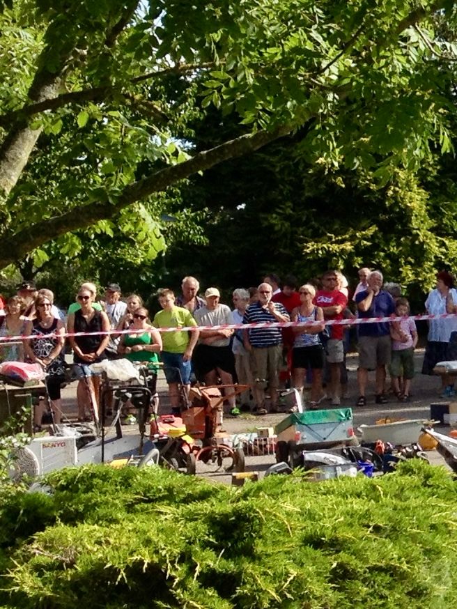 Das Bild zeigt eine Gruppe von Menschen, die im Freien hinter einem Absperrband stehen und offenbar auf etwas warten oder zuschauen. Im Vordergrund befindet sich eine Ansammlung verschiedener Gegenstände, die an einen Flohmarkt, eine Auktion oder einen Garagenverkauf erinnern. Darunter sind Kartons, Werkzeuge, kleinere Maschinen und weitere Alltagsgegenstände zu erkennen. Die Umgebung ist grün und von Bäumen sowie Büschen geprägt. Die Personen sind sommerlich gekleidet und das Wetter scheint sonnig zu sein. Die Atmosphäre wirkt belebt, und die Menschen stehen dicht beieinander, während sie das Geschehen beobachten.
