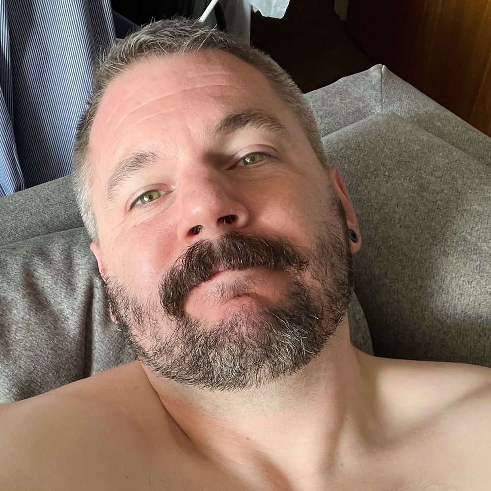 A close up of a man with short salt & pepper hair, green eyes, and a beard with a prominent mustache. He is looking up at the camera and appears to be topless.