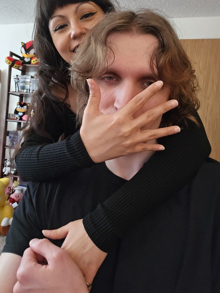 Picture of two people both in black clothes. One has their arms around the other's neck with their hand in front of face