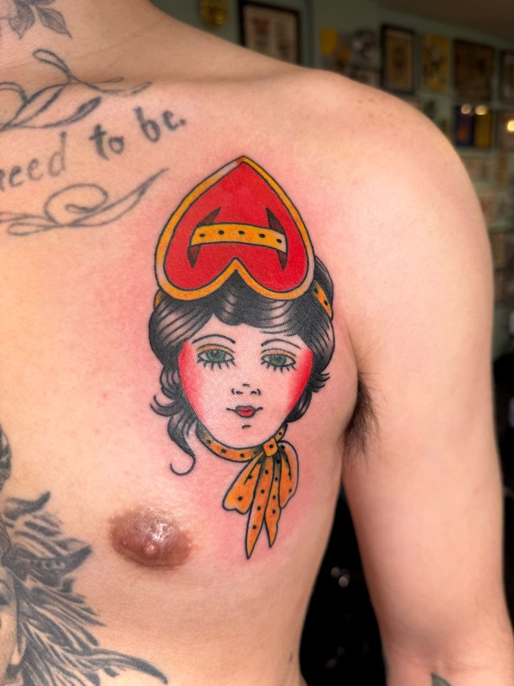 Lady head traditional color tattoo in the style of Bert Grimm with a heart head wrap. The piece is tattooed above a male’s left chest.