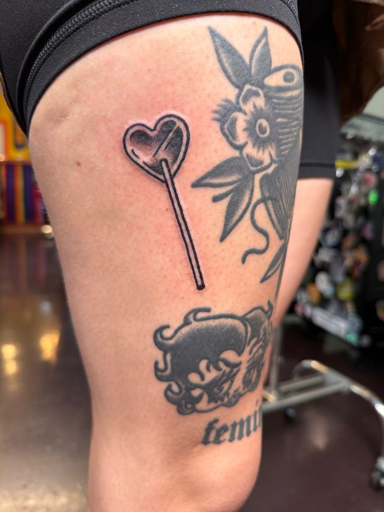 Black and grey traditional tattoo of a heart lollipop tattooed on the person’s thigh