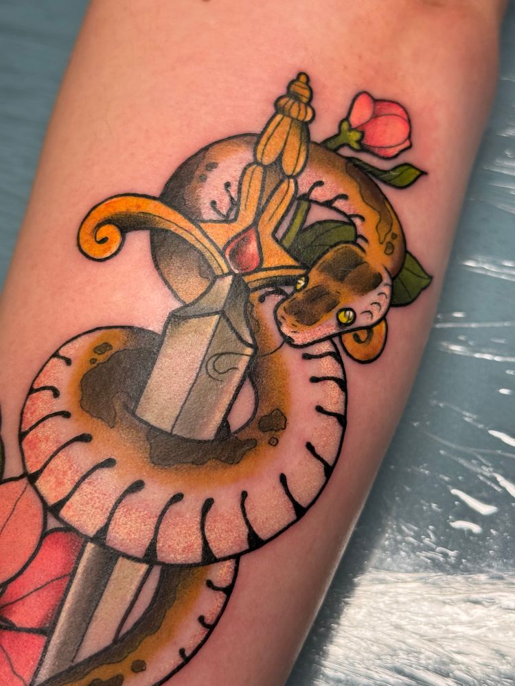 Close up — Photo of a fresh tattoo on the back of a forearm. The piece is a neo traditional snake wrapping around a dagger with a few flowers surrounding. The flowers are pink and the snake is a brown spotted color. You can also see the tongue sticking out and the bright green eyes.