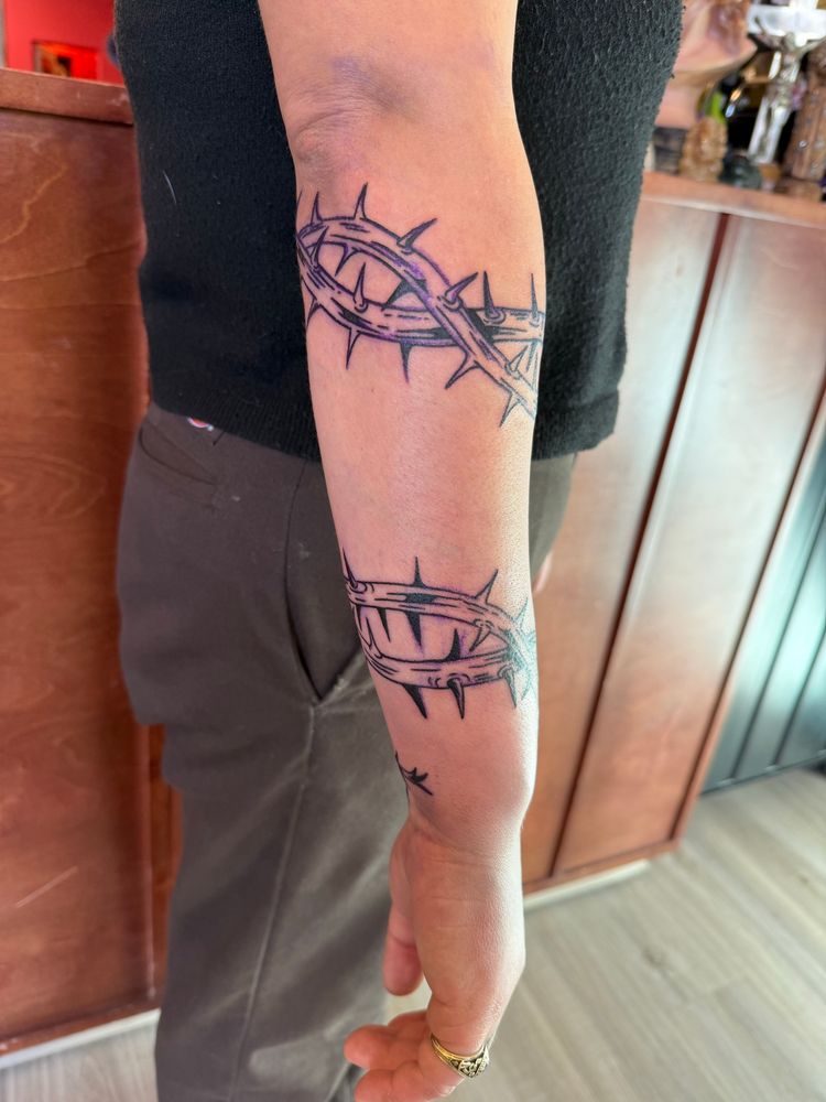 Side view. Photo of a light skinned person’s right arm with a fresh black work tattoo of a doubled thorny ocotillo branch wrapping around their forearm from their wrist to their elbow