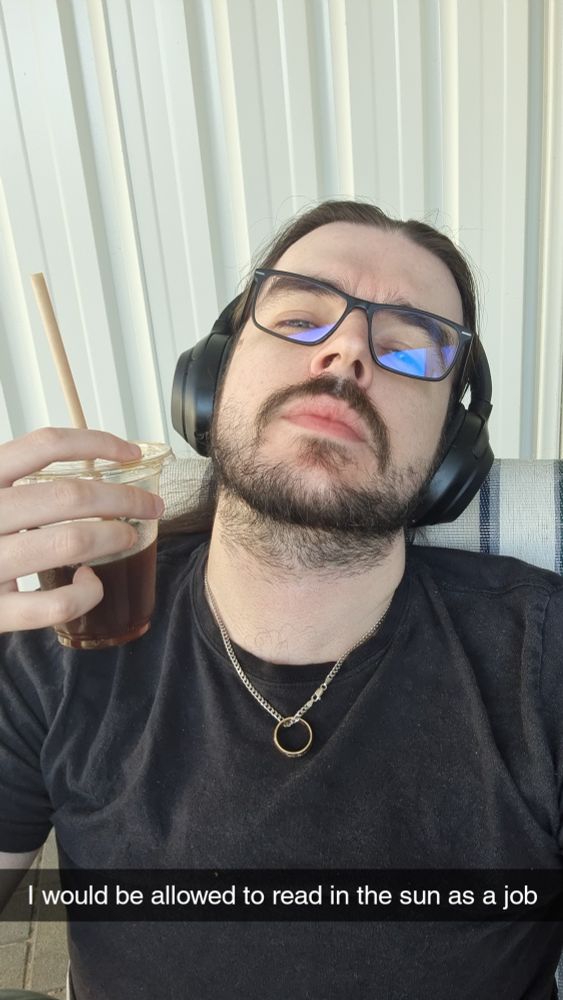 Riley is sitting outside looking very tired in a black shirt, wearing glasses and headphones while holding an iced coffee