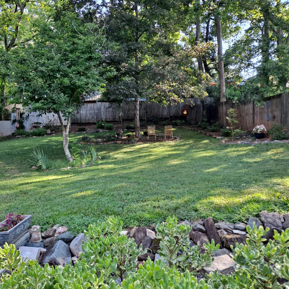 Early July scene. The magical little cottage's side garden with deep green lawn and maple tree. Two chairs sit waiting for you in the cool shade. Pots of colorful impatiens and caladiums are arranged around the garden. A lovely summer vignette. 