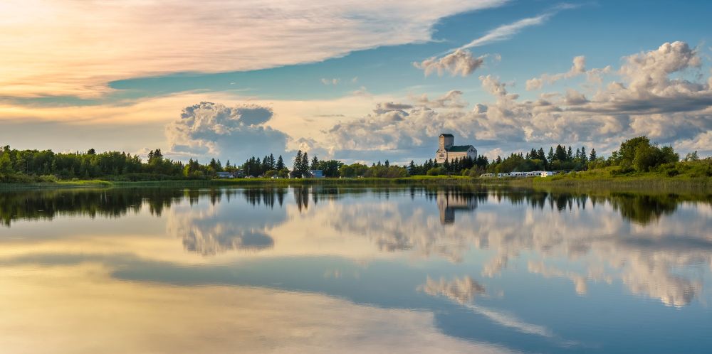Summer scene at Sandy Lake, Manitoba with a reflective lake showing a wide summer vista with clouds in the background. The town's grain elevator is shown as the town's prominent feature.