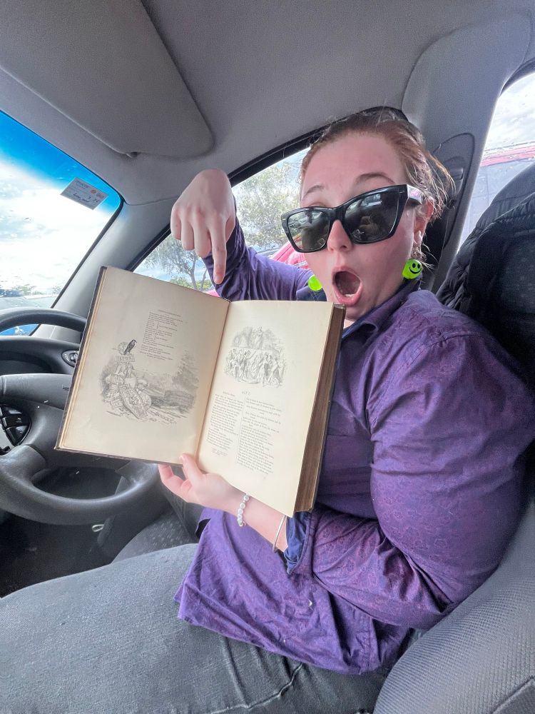 Me, same outfit as last photo. This time sitting in a car and excitedly showing one of the hand-drawn ink illustrations from my newly acquired collectible books to the camera. The camera is being held by my friend off screen. 