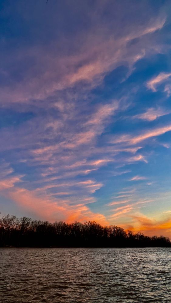 choppy water in a river flows by a darkened treeline on opposite riverbank. A sky with deep blue that fades to lighter shades is painted with wisps of pink above and orange below in this sunset photo.
