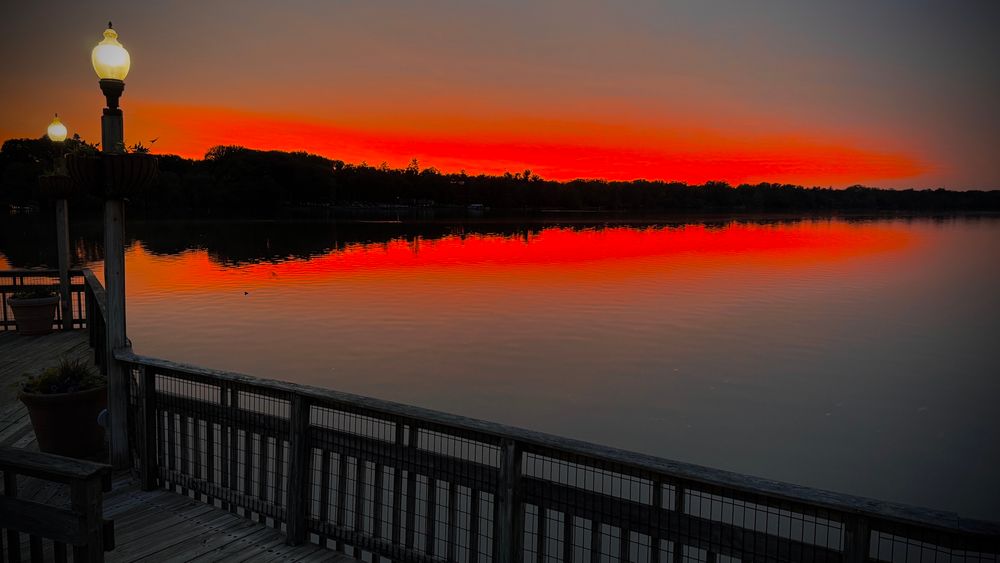 A crisp silhouette of a tree lined river bank in the distance contrasts sharply with a scarlet sky that fades to gray. The calm water mirrors the view. In foreground is a metal railing on a wooden boardwalk with illuminated  lamp posts and flower pots which runs from the lower right corner to middle of the left side of the sunset photo.