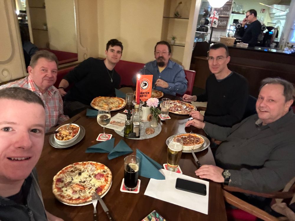 6 Personen sitzen an einem Tisch im Restaurant mit essen und trinken gedeckt. Alle schauen in die Kamera. 