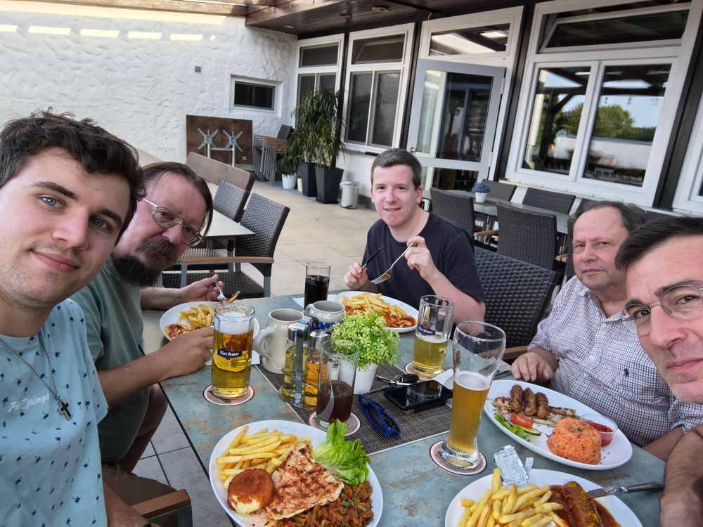 5 Personen sitzen in einer Gaststätte im Außenbereich an einem Tisch und haben Essen und Getränke vor sich stehen. Alle schauen in die Kamera. 