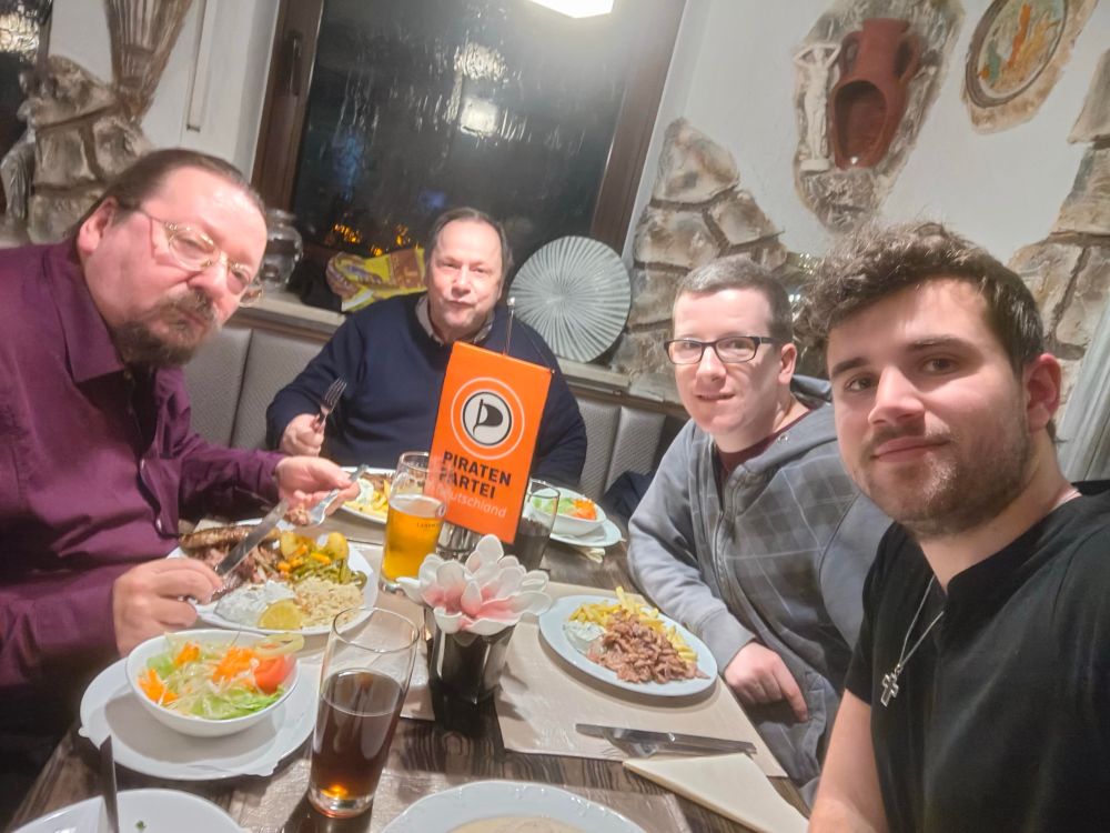 Vier Personen sitzen in einem Restaurant und haben Essen und Getränke vor sich. Sie schauen alle in die Kamera. 