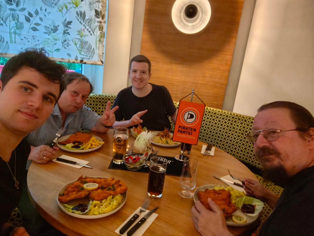 Vier Personen sitzen an einem runden Tisch und haben Spießen und Getränke vor sich! Sie schauen alle in die Kamera. Ein Piratenwimpel steht auf dem Tisch. 
