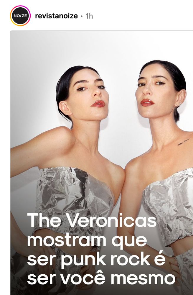 Duas mulheres idênticas, lado a lado, com cabelo preto liso preso para trás e batom vermelho. Elas usam vestidos prateados brilhantes que lembram papel alumínio. Estão de frente para a câmera com expressão séria. O fundo é branco e, na parte inferior da imagem, aparece o texto: “The Veronicas mostram que ser punk rock é ser você mesmo”.