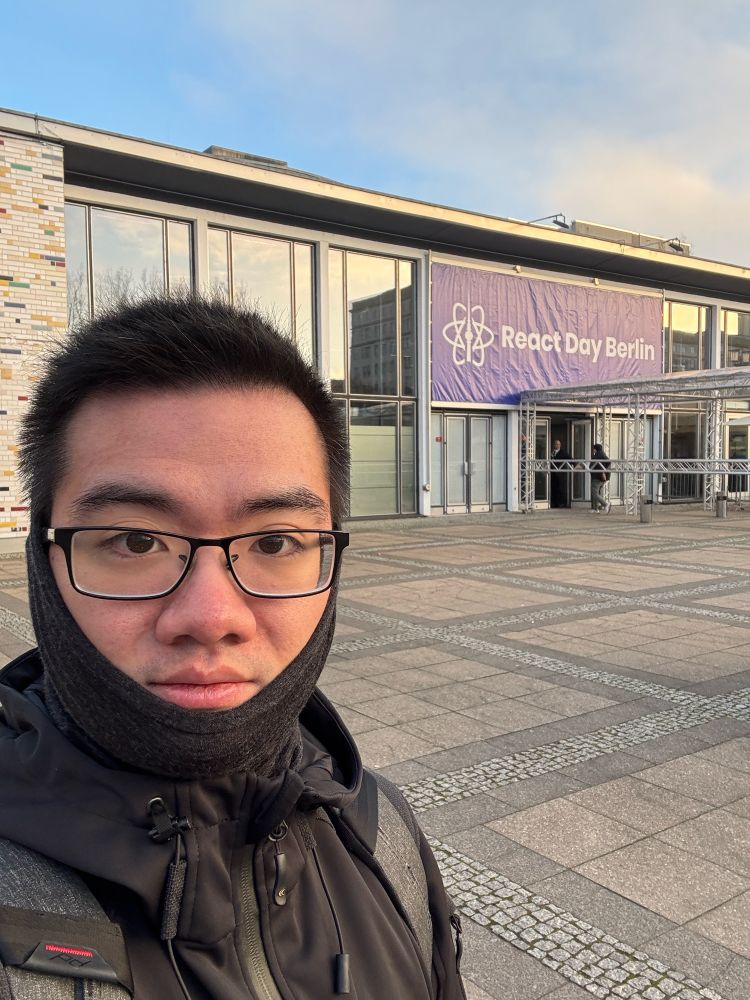 A person taking a selfie outside the entrance of React Day Berlin conference venue, wearing winter clothing. A purple banner with the React logo is visible on the building facade.