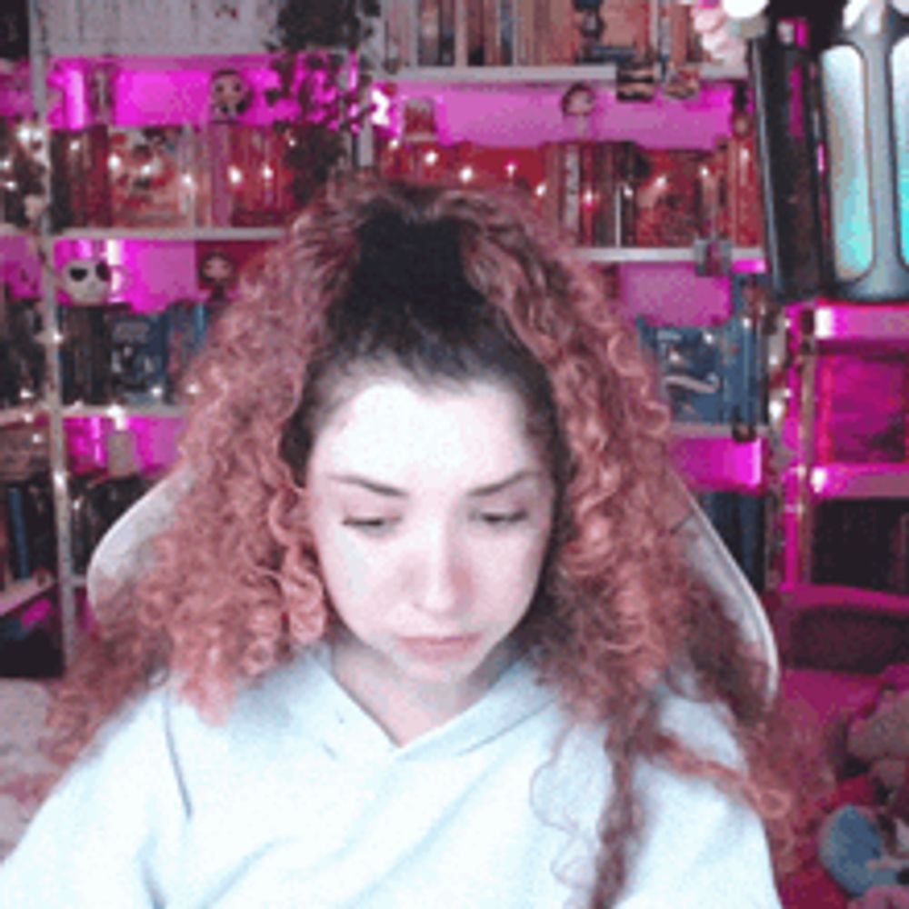 a girl with pink curly hair is sitting in front of a bookshelf with books on it .