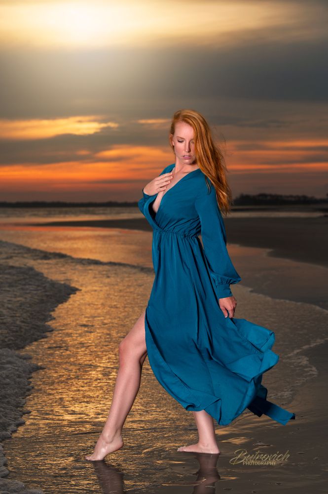 This striking photo captures a barefoot woman gracefully posing at the edge of the ocean during a vibrant sunset. She is wearing a flowing teal-blue dress that catches the breeze, adding a sense of motion and elegance to the image. Her red hair cascades over one shoulder, complementing the warm tones of the sky and the golden reflections in the wet sand beneath her. The background features a dramatic blend of deep oranges, purples, and soft grays in the sky, suggesting the sun has just dipped below the horizon. Her poised stance, with one leg extended and one hand resting gently on her chest, exudes confidence and serenity. The overall composition is both powerful and poetic, blending natural beauty with graceful movement.


