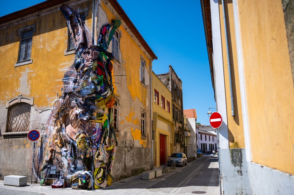 This photo captures a striking piece of urban street art on the side of a weathered yellow building by the artist Bordalo II. The artwork is a massive, three-dimensional mural of a rabbit, created using a combination of colorful paint and recycled materials such as scrap metal, plastic, and wood. The textures and objects give the rabbit a layered, sculptural quality that makes it appear to leap out from the wall. 

The building itself shows signs of age, with peeling paint and cracks, adding a sense of history and contrast to the vibrant modern artwork. A small cobblestone street runs alongside, lined with older buildings painted in muted tones, one with red doors and another in pale yellow. A car is parked further down the street, and traffic signs are visible — a "no entry" sign on the right wall and a "no parking" sign on the left near the artwork. Overhead, the sky is a brilliant clear blue, providing a vivid backdrop that enhances the bright colors of the mural.